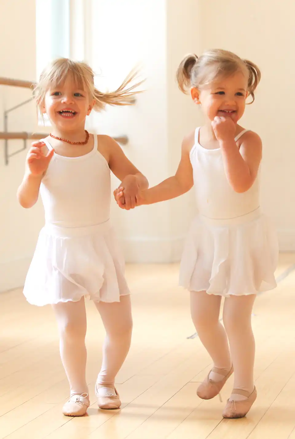 two smiling dancers in ballet uniforms holding hands in dance class